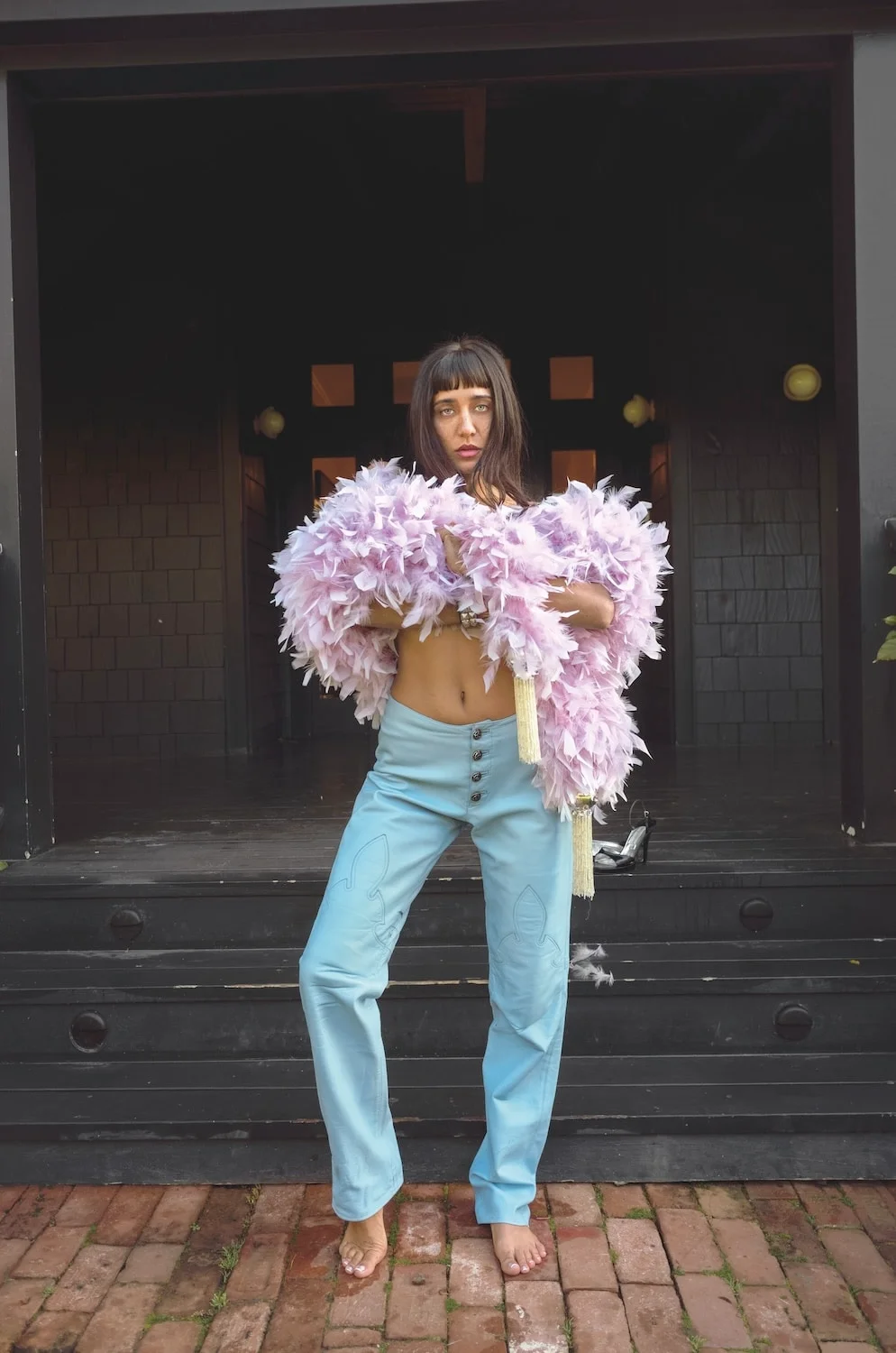 Jesse Jo Stark holding an armful of lilac feathers, wearing pale blue leather trousers, barefoot on a brick path in front of a black porch