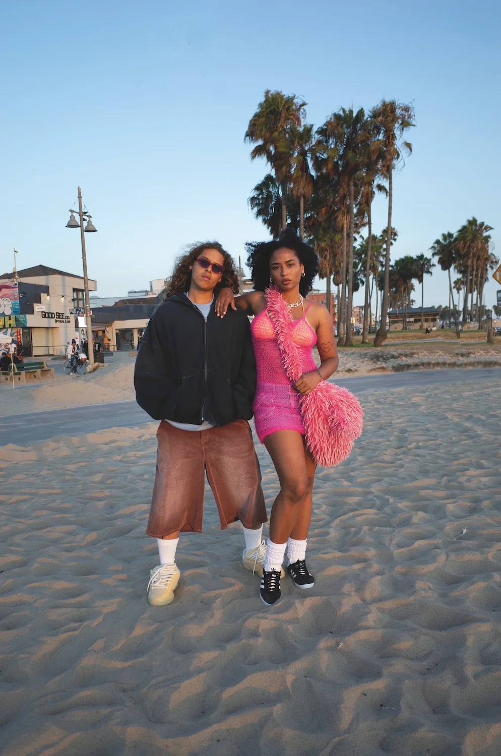 Two people standing on the sand at golden hour, one in an oversized black zip hoodie and brown corduroy shorts, the other in a pink crochet dress holding a shaggy pink bag, palm trees behind them